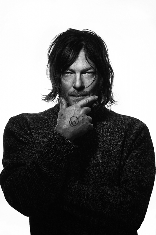 Black and white portrait of an adult male with medium-length hair, wearing a sweater and posing indoors with hand to chin.