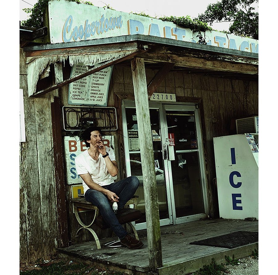 Adult male sitting on a bench smoking a cigarette outside of a small, weathered roadside store with an ice box and signage.