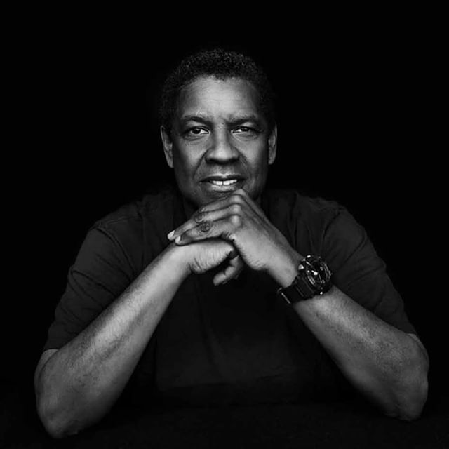 Monochrome portrait of adult man with hands clasped under his chin, wearing a dark t-shirt and wristwatch against a black background.