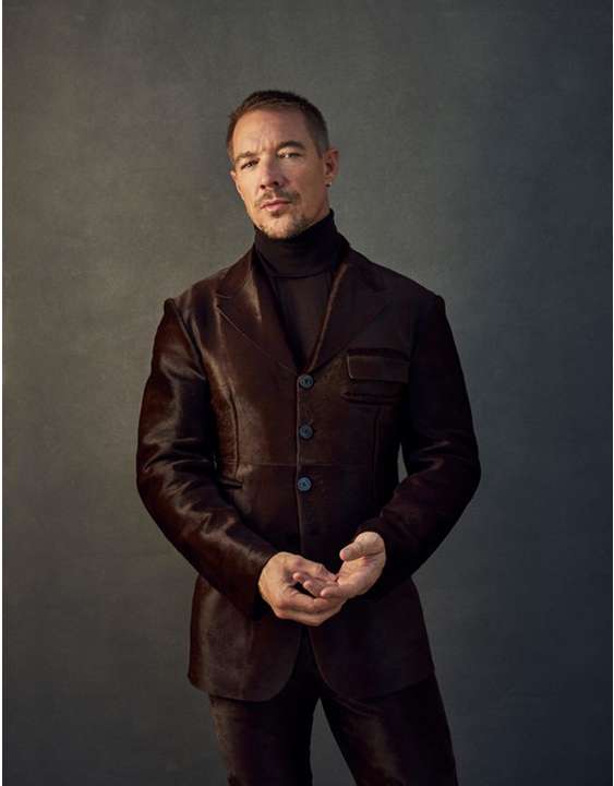 Portrait of an adult man wearing a brown suit and a black turtleneck against a neutral gray backdrop in a studio setting with soft lighting.