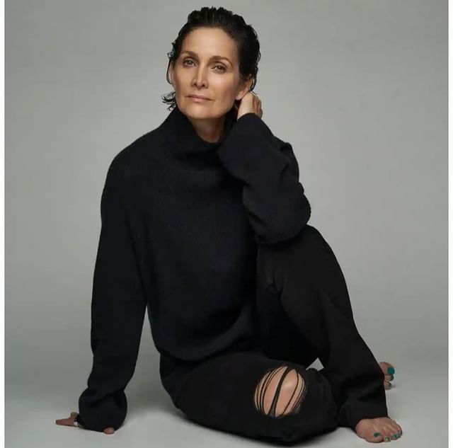 Carrie-Anne Moss seated wearing a black turtleneck sweater and black distressed jeans against a grey background.
