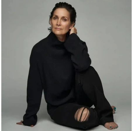 Carrie-Anne Moss seated wearing a black turtleneck sweater and black distressed jeans against a grey background.