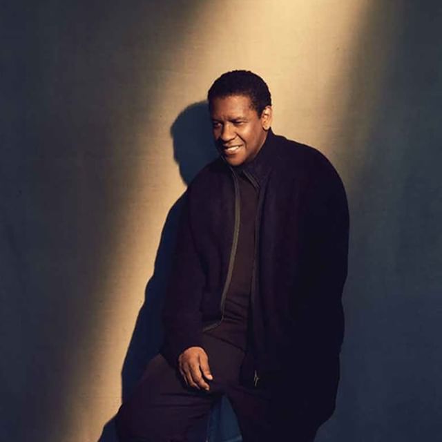 Portrait of Denzel Washington wearing a dark zip-up jacket against a neutral studio wall with a spotlight