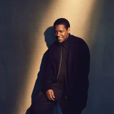 Portrait of Denzel Washington wearing a dark zip-up jacket against a neutral studio wall with a spotlight