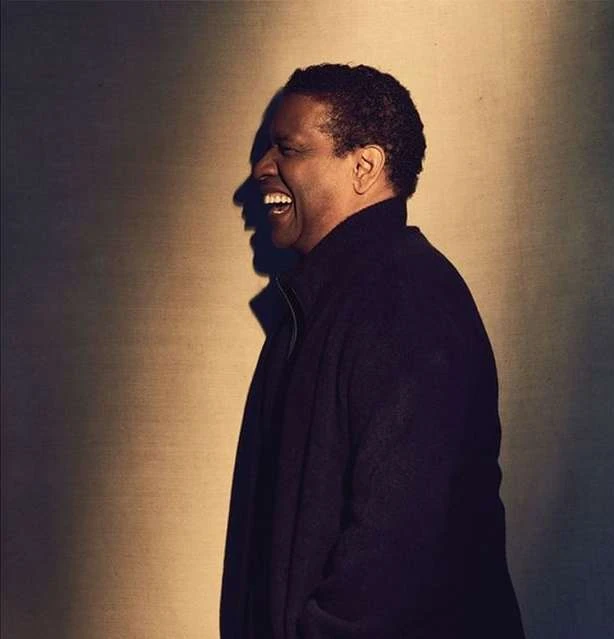 Denzel Washington in a medium studio shot, laughing in profile against a soft brown background. He wears a dark coat.