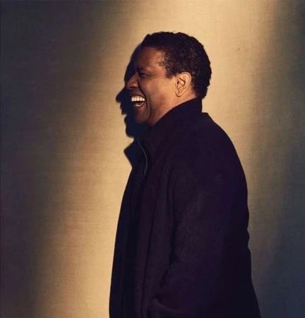 Denzel Washington in a medium studio shot, laughing in profile against a soft brown background. He wears a dark coat.