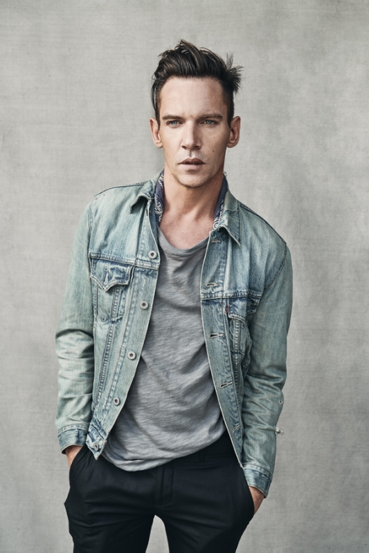 Portrait of a fair-skinned adult male in a gray t-shirt and denim jacket posing indoors against a neutral background in a studio shot.
