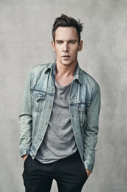 Portrait of a fair-skinned adult male in a gray t-shirt and denim jacket posing indoors against a neutral background in a studio shot.