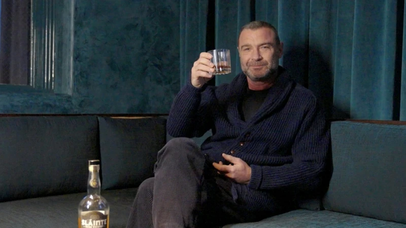 Portrait of adult man in a blue sweater raising a glass of amber liquor indoors on a couch next to a bottle of Irish liqueur.