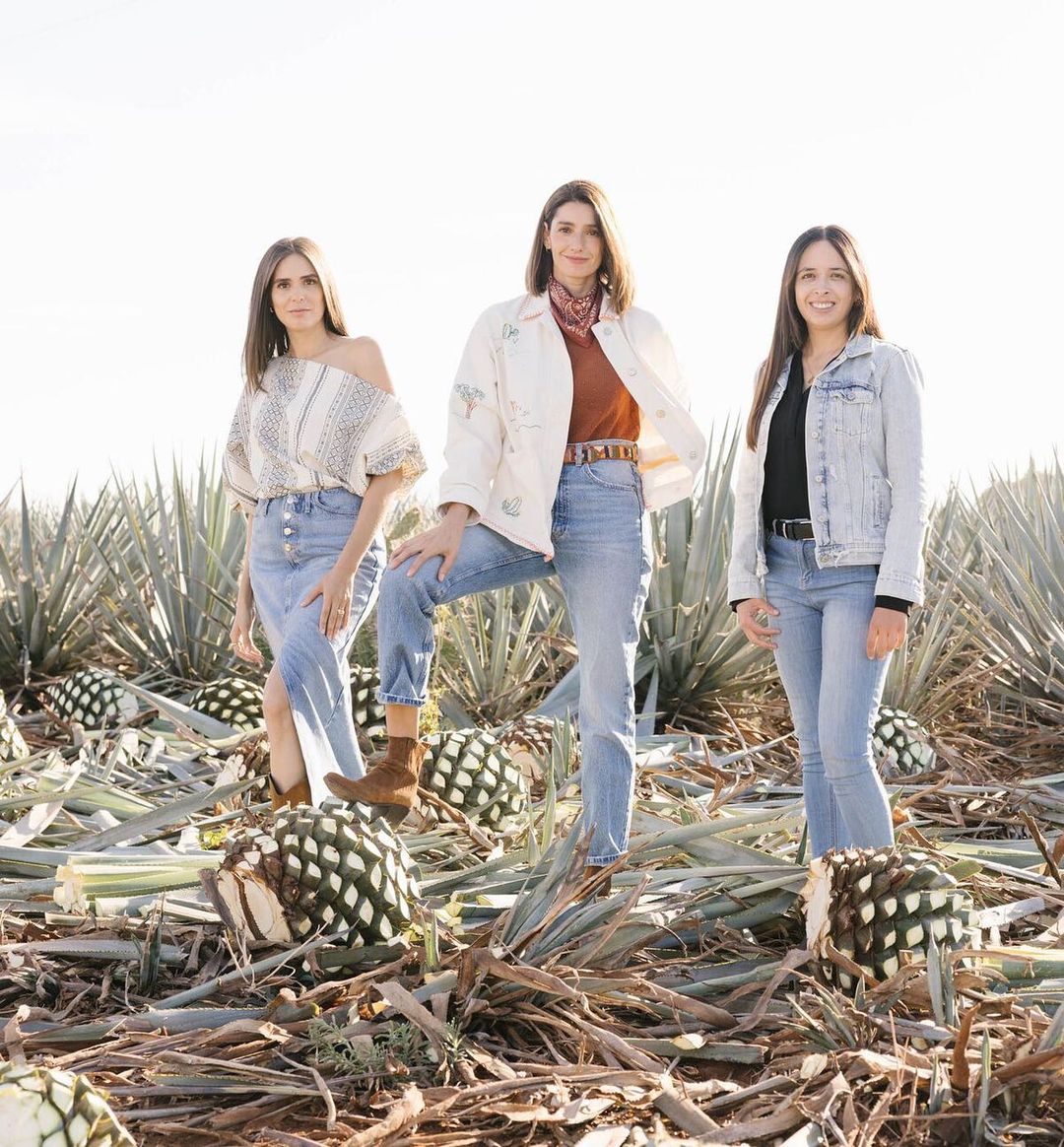 Three adult women in jeans pose near harvested agave plants in a rural field under bright daylight, wearing casual outerwear.