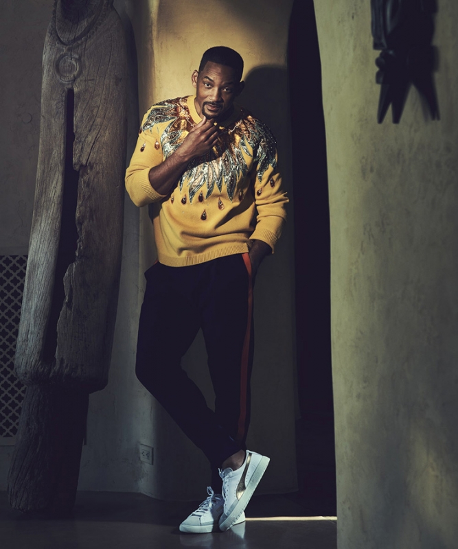 Portrait of Will Smith in a yellow patterned sweater, leaning against a wall in an indoor setting with decorative wood carving.