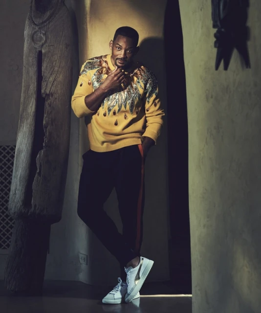 Portrait of Will Smith in a yellow patterned sweater, leaning against a wall in an indoor setting with decorative wood carving.