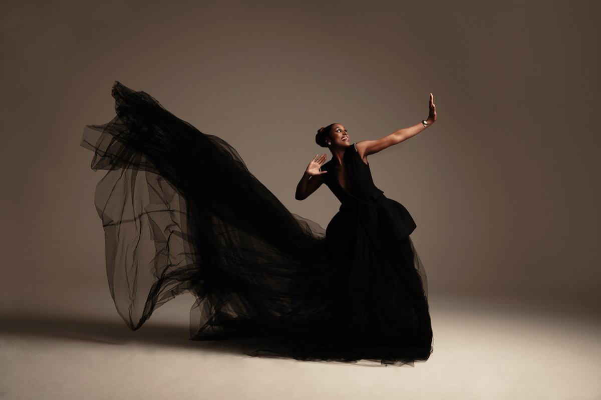 Woman in motion wearing black dress with long flowing train in studio. Arm raised with open palm. Lit from side. Neutral background.