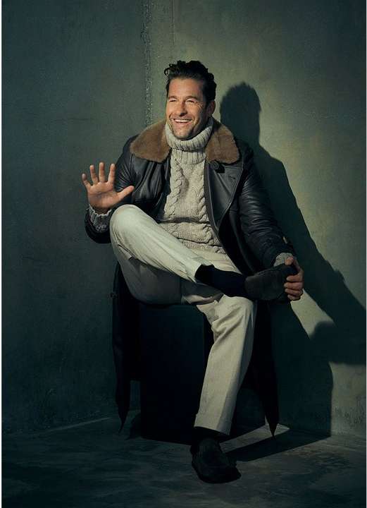 Portrait of an adult man in a jacket and roll neck sweater sitting, facing the camera and waving, in front of a grey background.