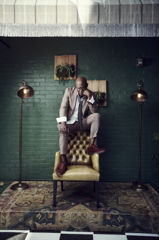 Bald man sits on a yellow chair indoors in a brown suit. He is resting his head on his hand, with two lamps behind him.