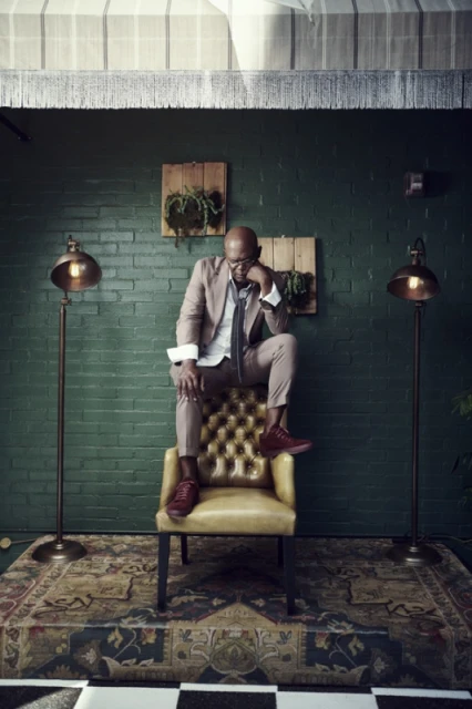 Bald man sits on a yellow chair indoors in a brown suit. He is resting his head on his hand, with two lamps behind him.