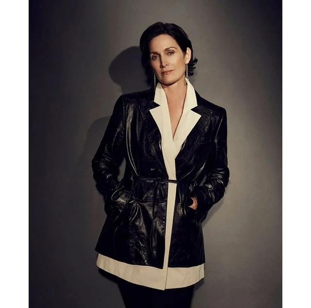 Portrait of Carrie-Anne Moss in a black and white blazer against a dark grey background, smiling gently at the camera.