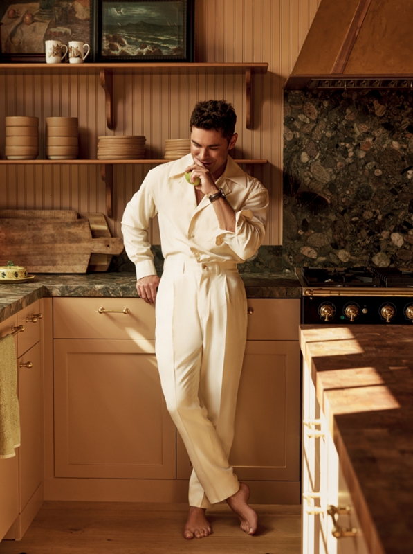 Barefoot adult male in a cream-colored suit eats green apple in a cream-colored kitchen with wood cabinets and a dark marble counter.
