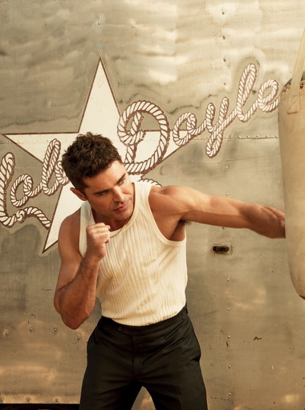 Adult male boxer in tank top and black pants in a fighting pose with a punching bag in a bright outdoor setting.
