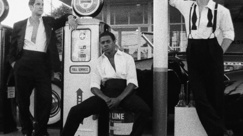 Three men in formal wear pose outside a gas station in a black and white photograph with vintage gas pumps and a car visible.