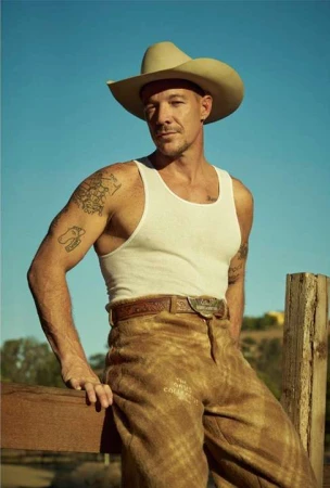 Man wearing a cowboy hat, white tank top, and patterned trousers resting his arm on a wooden fence against a clear sky background.