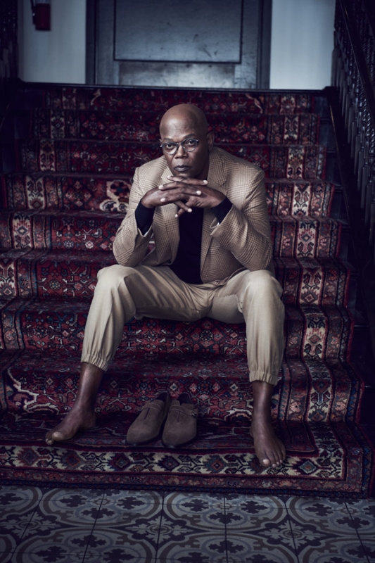 Samuel L. Jackson poses sitting on a staircase wearing a tan suit and no shoes, with his hands clasped, gazing at the viewer.