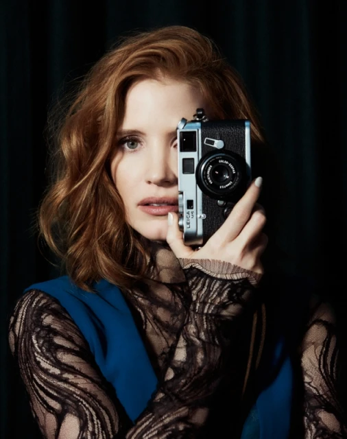 Portrait of red-haired woman with camera partially obscuring her face; she's wearing black lace long sleeves and a blue vest.