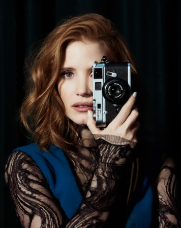 Portrait of red-haired woman with camera partially obscuring her face; she's wearing black lace long sleeves and a blue vest.
