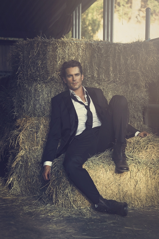 Adult male model wearing a suit posing on hay bales in an indoor barn setting with natural lighting and a medium camera shot.