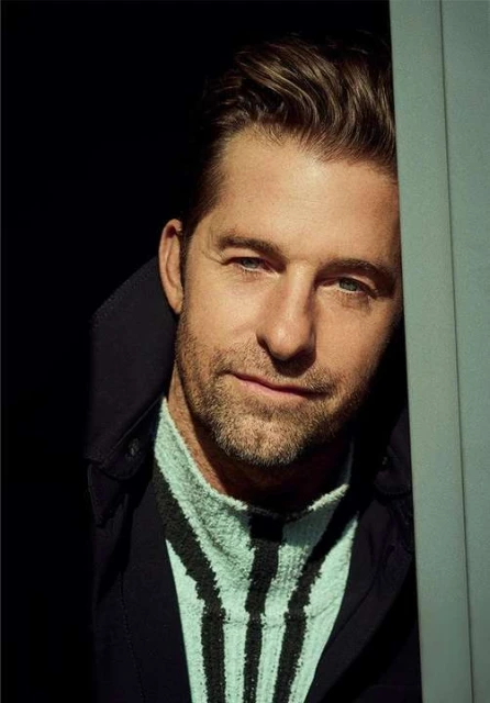 Portrait of a man looking at the camera with a slight smile wearing a navy jacket and striped sweater in front of a black background.