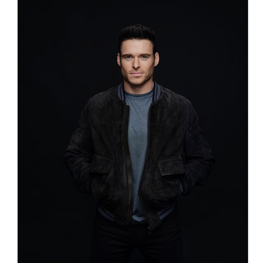 Portrait of adult male wearing a suede bomber jacket and T-shirt posing in front of a dark backdrop in a photo studio.