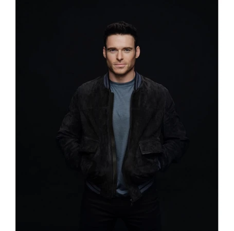 Portrait of adult male wearing a suede bomber jacket and T-shirt posing in front of a dark backdrop in a photo studio.