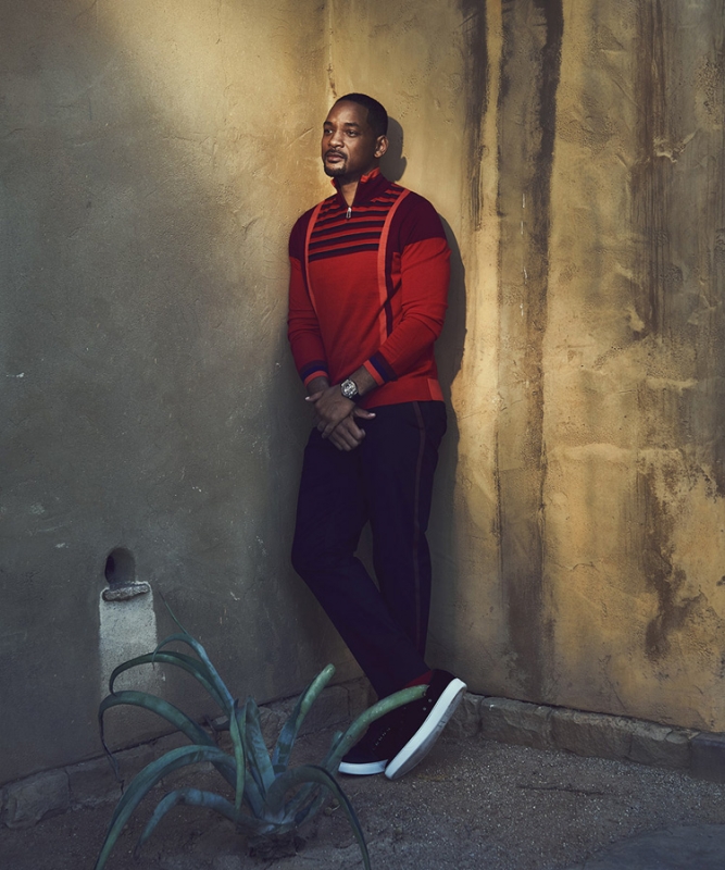 Portrait of an adult male in a red shirt and black pants leaning against a textured wall with a small plant in the corner.