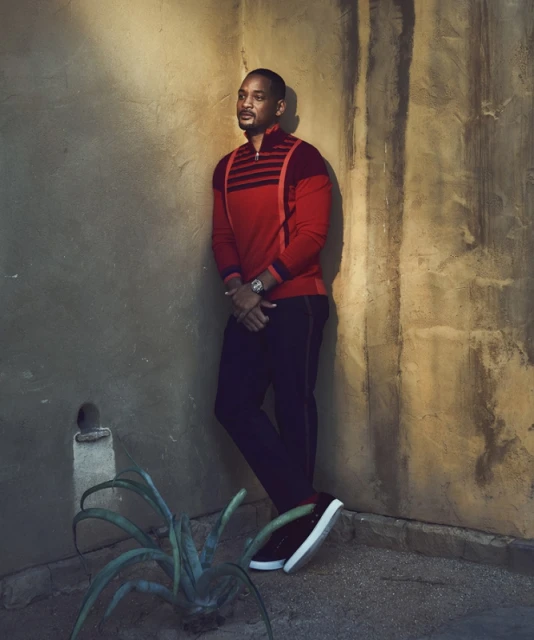 Portrait of an adult male in a red shirt and black pants leaning against a textured wall with a small plant in the corner.