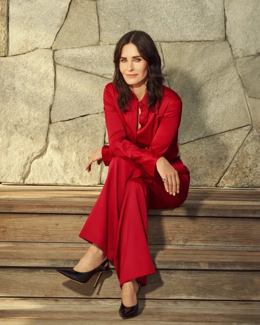 Courteney Cox, an adult female, sits on wooden stairs wearing a red pantsuit with black high heels against a stone wall backdrop.