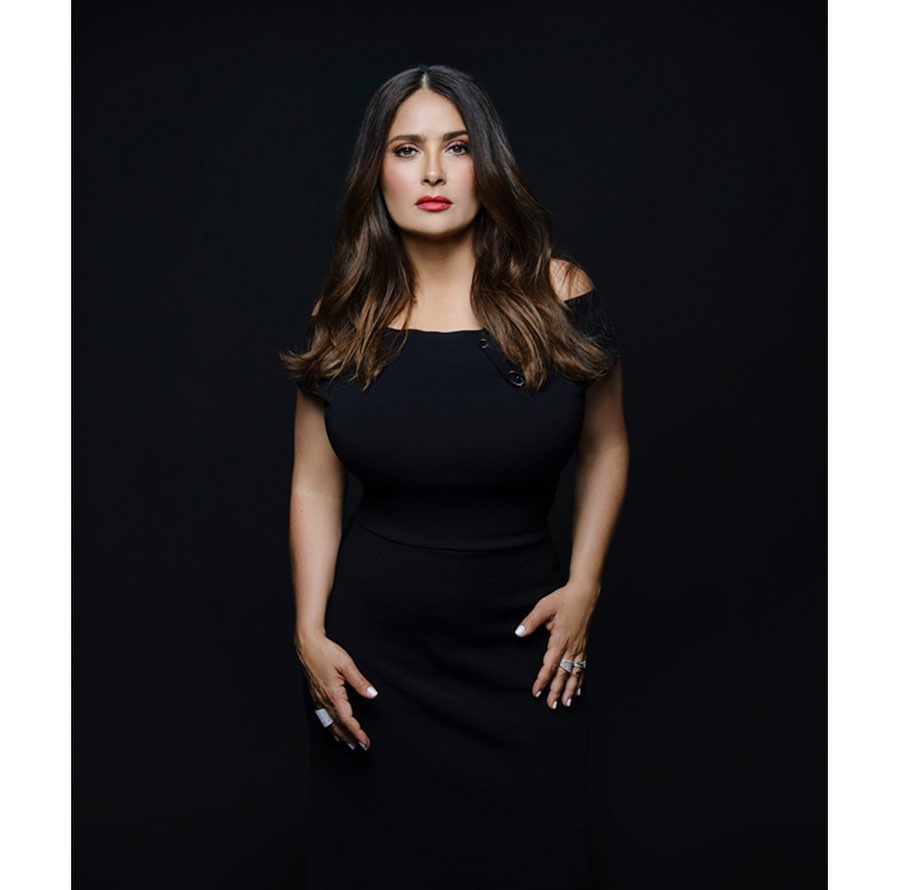 Salma Hayek poses in a black off-the-shoulder dress against a dark background. The portrait showcases her brown hair and red lipstick.