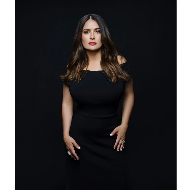 Salma Hayek poses in a black off-the-shoulder dress against a dark background. The portrait showcases her brown hair and red lipstick.