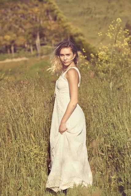 Woman in a white dress poses in a field of tall grass on a sunny day, with a soft, dreamy aesthetic and natural backdrop.