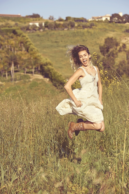 Young woman in sundress and cowboy boots jumps for joy in a grassy field on a sunny day. Rolling hills and blue sky background.