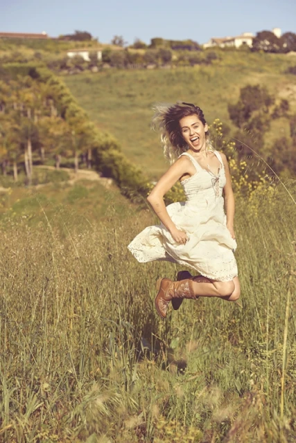 Young woman in sundress and cowboy boots jumps for joy in a grassy field on a sunny day. Rolling hills and blue sky background.
