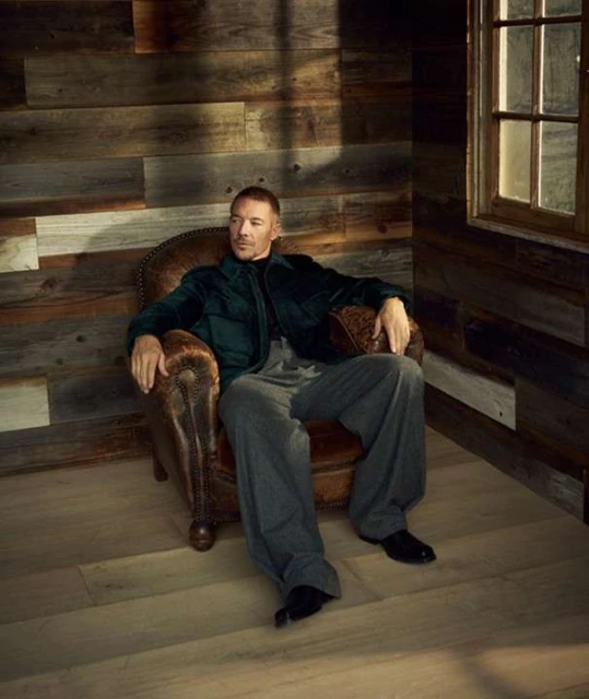 An adult male in a green jacket and gray pants sits in a leather armchair in a room with wood-paneled walls near a window with natural light.