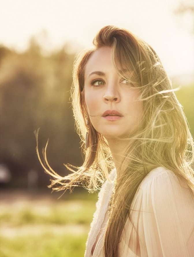 Portrait of a blonde adult woman in an outdoor setting, with sunlight highlighting her hair and a soft, blurred background of green foliage.