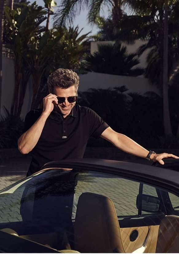 Man wearing sunglasses and a black polo shirt leaning against a car exterior outdoors