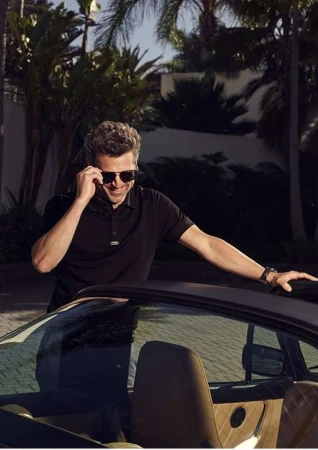 Man wearing sunglasses and a black polo shirt leaning against a car exterior outdoors