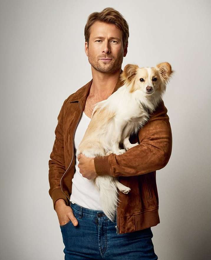 Blond adult male in a brown suede jacket holds a tan and white dog in a medium shot against a white background in a studio.