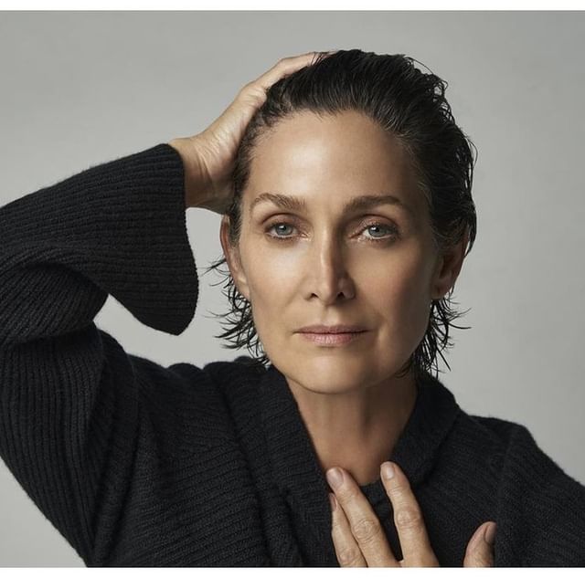 Portrait of a mature adult woman with wet, dark hair, wearing a black sweater, looking directly at the camera in a photo studio setting.