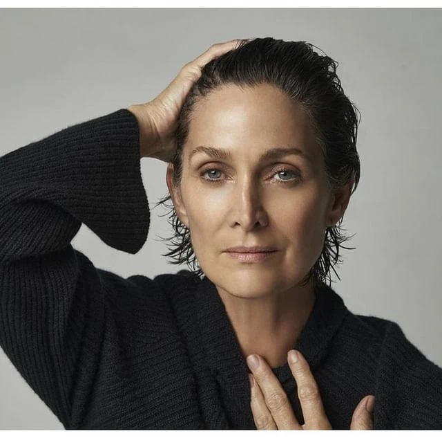 Portrait of a mature adult woman with wet, dark hair, wearing a black sweater, looking directly at the camera in a photo studio setting.
