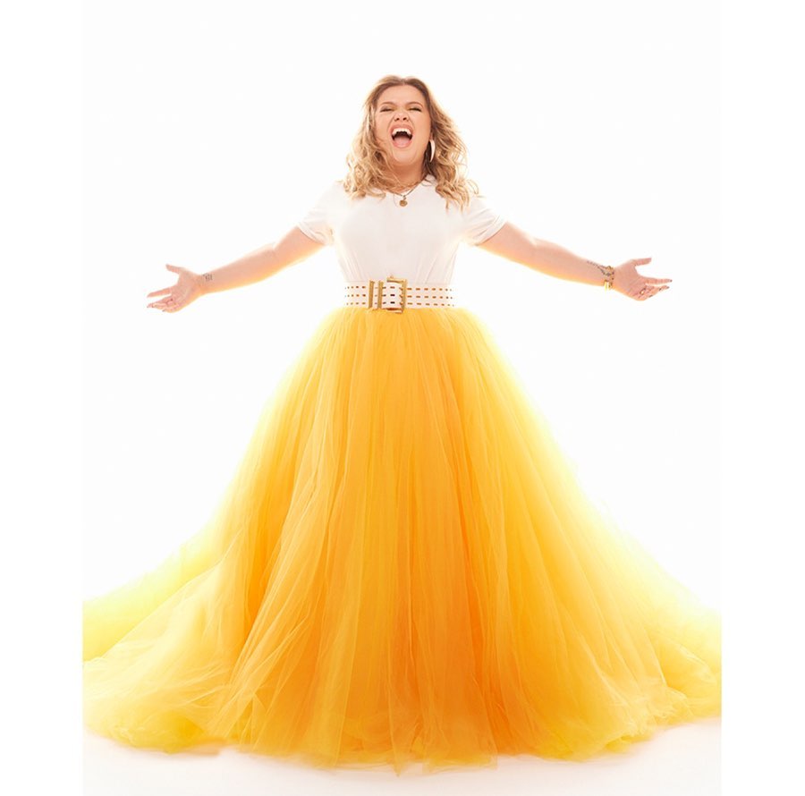Woman in white top and yellow tulle gown poses with arms outstretched against a bright white backdrop, conveying joyful energy.
