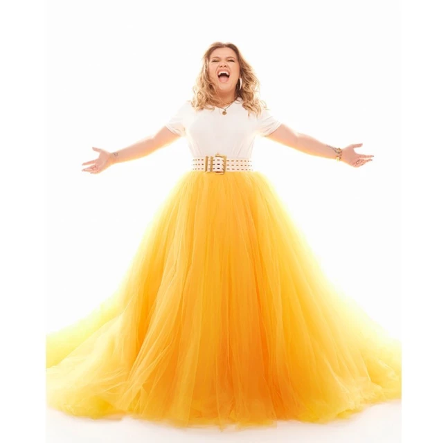 Woman in white top and yellow tulle gown poses with arms outstretched against a bright white backdrop, conveying joyful energy.