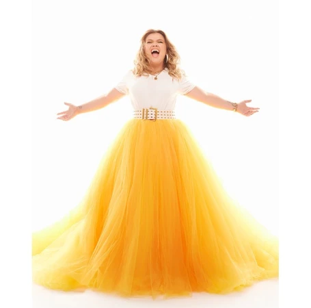 Woman in white top and yellow tulle gown poses with arms outstretched against a bright white backdrop, conveying joyful energy.